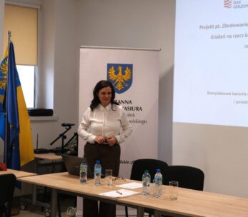 Portretowe ujęcie Wicemarszałek Zuzanny Donath-Kasiury stojącej przy ekranie z prezentacją projektu „Instytutu Śląskiego”. Prelegentka ubrana jest w białą koszulę i brązowe spodnie, a jej postawa sugeruje aktywny dialog z uczestnikami. Na stole przed nią stoją butelki z wodą mineralną i laptop.
