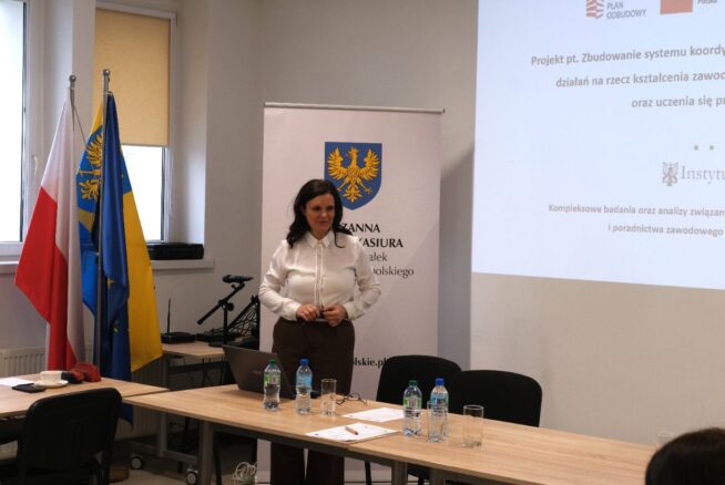 Portretowe ujęcie Wicemarszałek Zuzanny Donath-Kasiury stojącej przy ekranie z prezentacją projektu „Instytutu Śląskiego”. Prelegentka ubrana jest w białą koszulę i brązowe spodnie, a jej postawa sugeruje aktywny dialog z uczestnikami. Na stole przed nią stoją butelki z wodą mineralną i laptop.