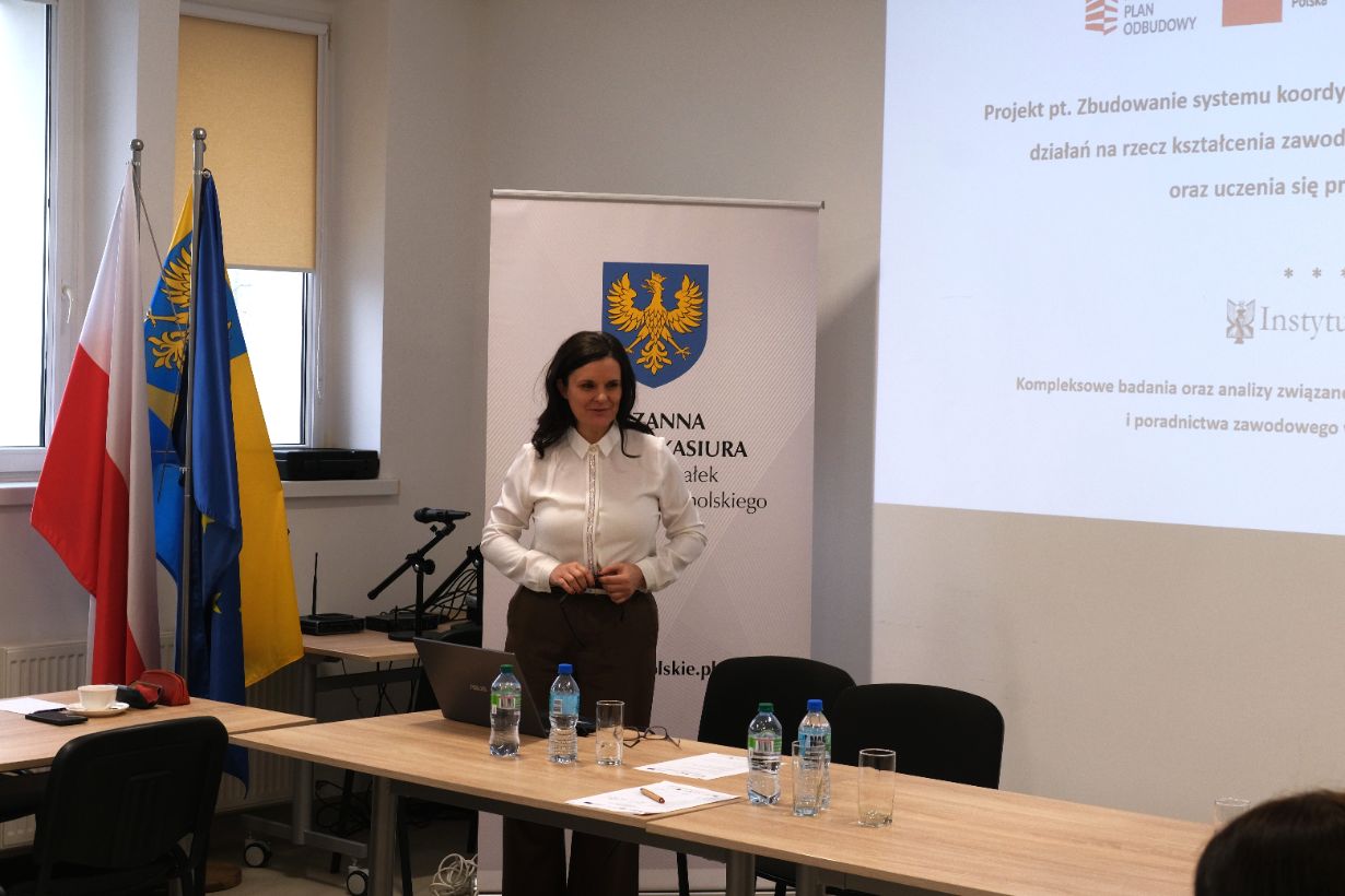 Portretowe ujęcie Wicemarszałek Zuzanny Donath-Kasiury stojącej przy ekranie z prezentacją projektu „Instytutu Śląskiego”. Prelegentka ubrana jest w białą koszulę i brązowe spodnie, a jej postawa sugeruje aktywny dialog z uczestnikami. Na stole przed nią stoją butelki z wodą mineralną i laptop.