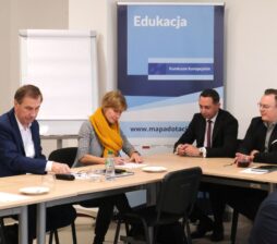 Cztery osoby siedzą przy stole konferencyjnym pod banerem „Edukacja – Fundusze Europejskie”. Kobieta w żółtym, grubym szalu notuje coś na dokumentach, podczas gdy mężczyźni obok niej obserwują przebieg spotkania. Po lewej stronie widać fragment tablicy z mapą i opisem regionu. Jest to reprezentacyjne ujęcie dokumentujące współpracę instytucjonalną.