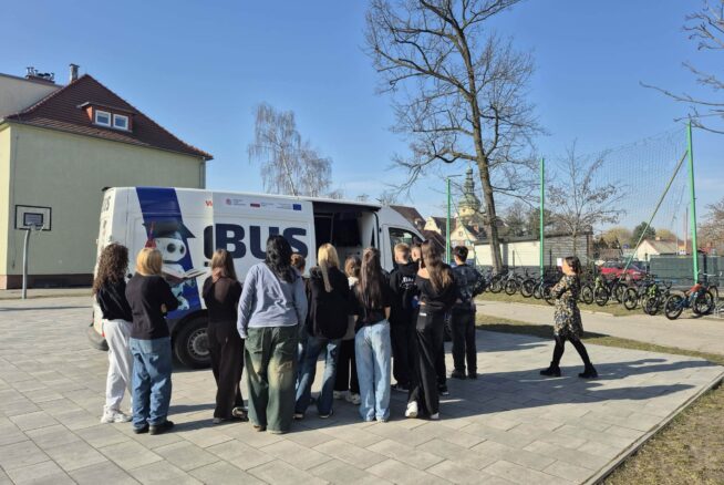 Na brukowanym placu przed busem z otwartymi drzwiami stoi grupa młodzieży i dorośli, oglądając wnętrze pojazdu. W tle widać budynek szkoły, drzewa i stojaki na rowery pod czystym, błękitnym niebem.