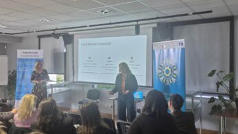 Zdjęcie przedstawia prelegentkę stojącą obok ekranu z napisem „CELE MEDIACJI SZKOLNEJ” podczas spotkania w nowoczesnej sali konferencyjnej. Uczestnicy siedzą tyłem do obiektywu, słuchając wystąpienia, a obok ekranu widoczne są banery informacyjne, w tym jeden z logo Powiatu Strzeleckiego. Pomieszczenie jest jasne, z podwieszanym sufitem i drewnianą podłogą.