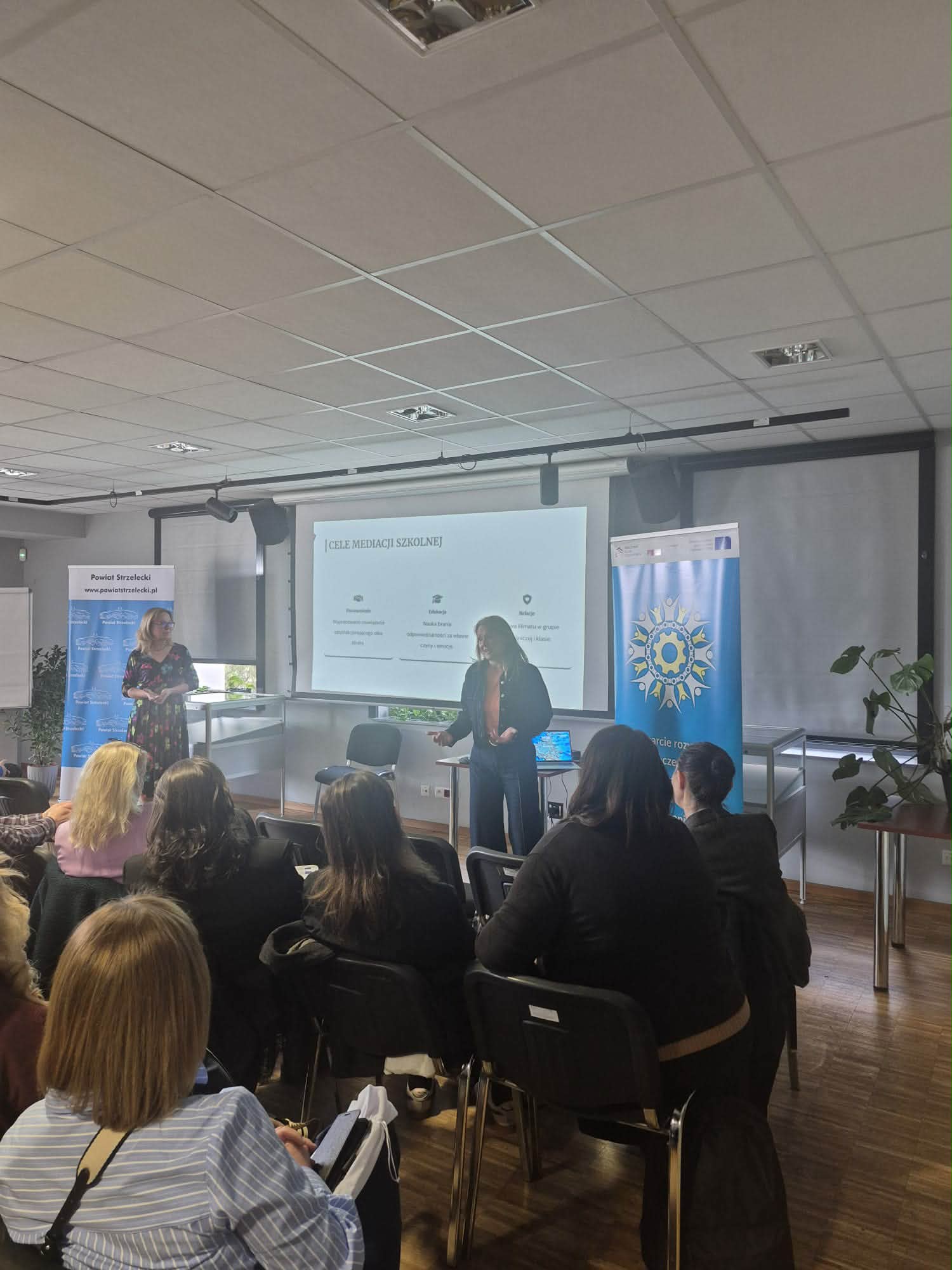 Zdjęcie przedstawia prelegentkę stojącą obok ekranu z napisem „CELE MEDIACJI SZKOLNEJ” podczas spotkania w nowoczesnej sali konferencyjnej. Uczestnicy siedzą tyłem do obiektywu, słuchając wystąpienia, a obok ekranu widoczne są banery informacyjne, w tym jeden z logo Powiatu Strzeleckiego. Pomieszczenie jest jasne, z podwieszanym sufitem i drewnianą podłogą.