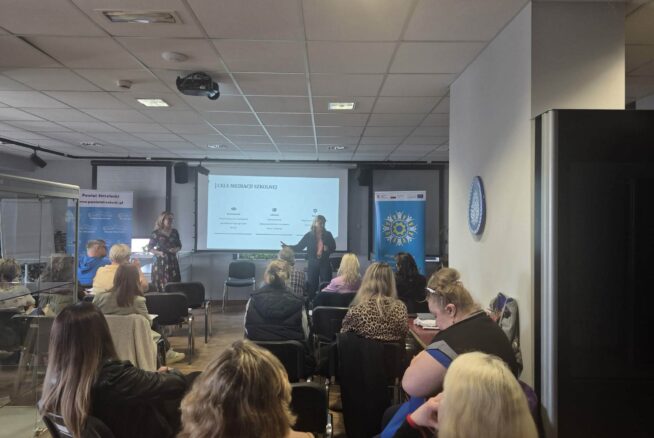 Szerokie ujęcie sali konferencyjnej wypełnionej osobami biorącymi udział w szkoleniu na temat mediacji szkolnej. Dwie kobiety prowadzą prezentację przy ekranie multimedialnym, a słuchacze siedzą w rzędach krzeseł, wypełniając niemal całą dostępną przestrzeń. Wnętrze jest profesjonalnie wyposażone, z widocznym rzutnikiem pod sufitem i dekoracyjnymi elementami na ścianach.