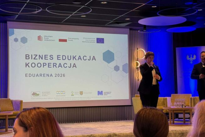 Kobieta w ciemnym garniturze przemawia do mikrofonu na scenie podczas konferencji „EduArena 2026”. W tle widoczny jest duży ekran z napisami „Biznes Edukacja Kooperacja” oraz logotypami instytucji partnerskich i Unii Europejskiej. Po prawej stronie sceny znajduje się fragment sylwetki mężczyzny oraz nowoczesne fotele i stolik z wodą.