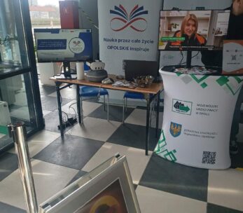 Stanowisko Wojewódzkiego Urzędu Pracy w Opolu z banerem pod hasłem „Nauka przez całe życie – OPOLSKIE inspiruje”. Na biurku przygotowano zestaw VR do wirtualnego poznawania zawodów, a na monitorach wyświetlane są materiały informacyjne. Obok stoi biały stand z logo urzędu oraz kodem QR.