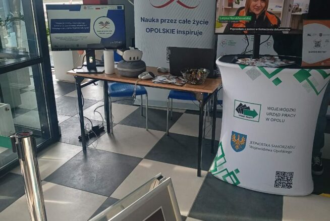 Stanowisko Wojewódzkiego Urzędu Pracy w Opolu z banerem pod hasłem „Nauka przez całe życie – OPOLSKIE inspiruje”. Na biurku przygotowano zestaw VR do wirtualnego poznawania zawodów, a na monitorach wyświetlane są materiały informacyjne. Obok stoi biały stand z logo urzędu oraz kodem QR.