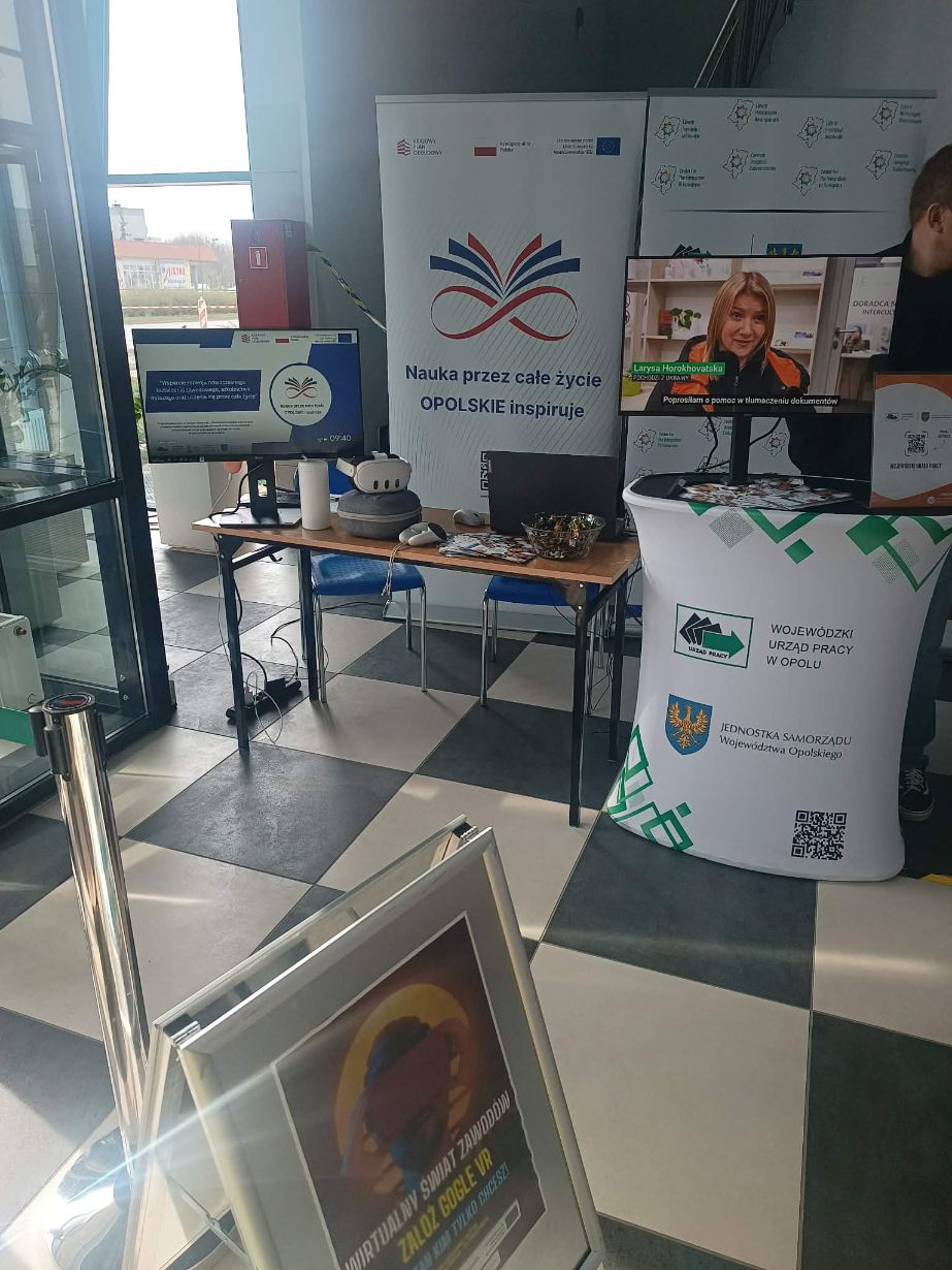 Stanowisko Wojewódzkiego Urzędu Pracy w Opolu z banerem pod hasłem „Nauka przez całe życie – OPOLSKIE inspiruje”. Na biurku przygotowano zestaw VR do wirtualnego poznawania zawodów, a na monitorach wyświetlane są materiały informacyjne. Obok stoi biały stand z logo urzędu oraz kodem QR.