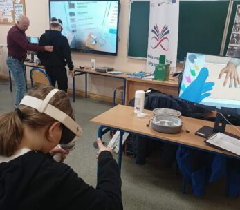 Grupa osób w sali lekcyjnej podczas warsztatów z wykorzystaniem nowoczesnych technologii. Na pierwszym planie dziewczynka w goglach VR obsługuje kontrolery, a w tle widać duży ekran interaktywny z listą zawodów oraz baner promocyjny. Instruktor w bordowym swetrze asystuje innemu uczniowi przy stanowisku komputerowym.