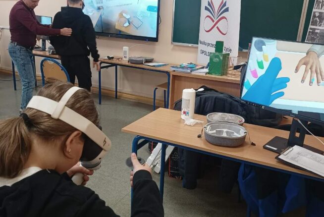 Grupa osób w sali lekcyjnej podczas warsztatów z wykorzystaniem nowoczesnych technologii. Na pierwszym planie dziewczynka w goglach VR obsługuje kontrolery, a w tle widać duży ekran interaktywny z listą zawodów oraz baner promocyjny. Instruktor w bordowym swetrze asystuje innemu uczniowi przy stanowisku komputerowym.