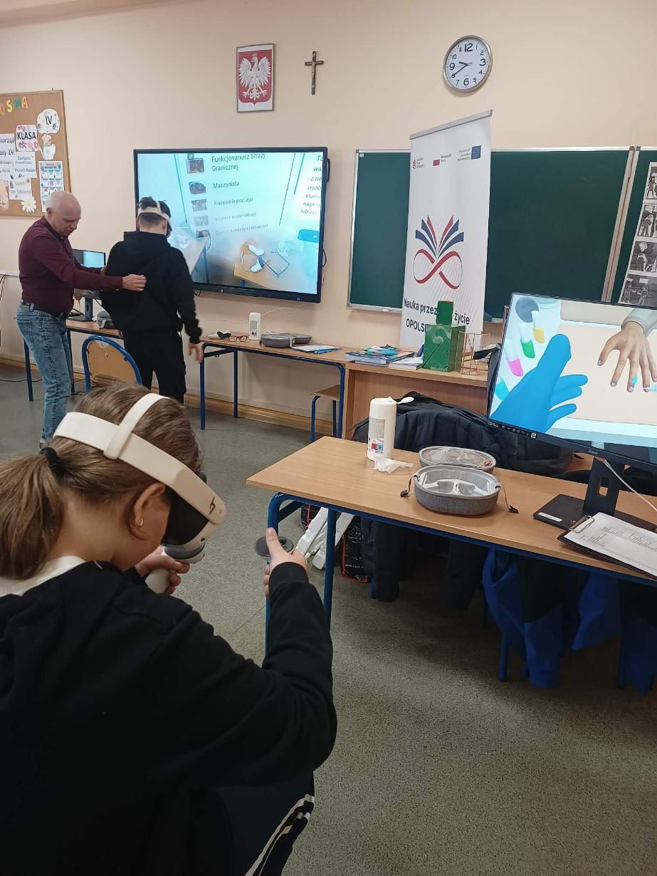 Grupa osób w sali lekcyjnej podczas warsztatów z wykorzystaniem nowoczesnych technologii. Na pierwszym planie dziewczynka w goglach VR obsługuje kontrolery, a w tle widać duży ekran interaktywny z listą zawodów oraz baner promocyjny. Instruktor w bordowym swetrze asystuje innemu uczniowi przy stanowisku komputerowym.