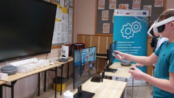 Na zdjęciu widać młodego mężczyznę w turkusowej koszulce, który używa gogli VR i dwóch kontrolerów do obsługi interaktywnego programu. Obraz z jego gogli, przedstawiający warsztat z narzędziami, jest dublowany na monitorze stojącym na biurku przed nim. W tle stoi niebieski baner informacyjny z logotypami Unii Europejskiej oraz grafiką kół zębatych. Sala wyposażona jest w nowoczesny sprzęt edukacyjny, w tym duży ekran dotykowy zamontowany na ścianie.
