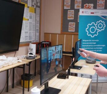 Na zdjęciu widać młodego mężczyznę w turkusowej koszulce, który używa gogli VR i dwóch kontrolerów do obsługi interaktywnego programu. Obraz z jego gogli, przedstawiający warsztat z narzędziami, jest dublowany na monitorze stojącym na biurku przed nim. W tle stoi niebieski baner informacyjny z logotypami Unii Europejskiej oraz grafiką kół zębatych. Sala wyposażona jest w nowoczesny sprzęt edukacyjny, w tym duży ekran dotykowy zamontowany na ścianie.
