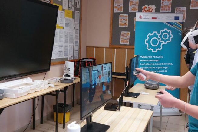 Na zdjęciu widać młodego mężczyznę w turkusowej koszulce, który używa gogli VR i dwóch kontrolerów do obsługi interaktywnego programu. Obraz z jego gogli, przedstawiający warsztat z narzędziami, jest dublowany na monitorze stojącym na biurku przed nim. W tle stoi niebieski baner informacyjny z logotypami Unii Europejskiej oraz grafiką kół zębatych. Sala wyposażona jest w nowoczesny sprzęt edukacyjny, w tym duży ekran dotykowy zamontowany na ścianie.