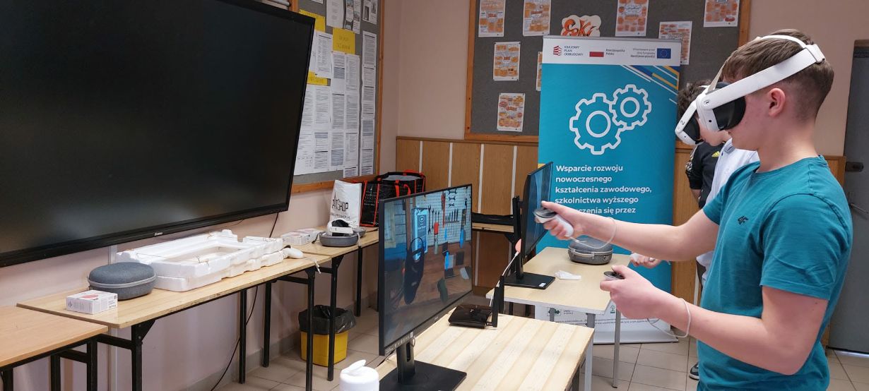 Na zdjęciu widać młodego mężczyznę w turkusowej koszulce, który używa gogli VR i dwóch kontrolerów do obsługi interaktywnego programu. Obraz z jego gogli, przedstawiający warsztat z narzędziami, jest dublowany na monitorze stojącym na biurku przed nim. W tle stoi niebieski baner informacyjny z logotypami Unii Europejskiej oraz grafiką kół zębatych. Sala wyposażona jest w nowoczesny sprzęt edukacyjny, w tym duży ekran dotykowy zamontowany na ścianie.