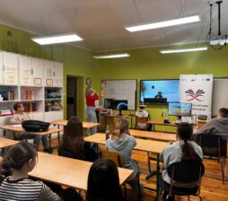 Zdjęcie ukazuje grupę uczniów siedzących w ławkach i patrzących w stronę tablicy multimedialnej, na której wyświetlane jest połączenie wideo. W centralnej części sali dwoje uczniów korzysta z zestawów VR, a nauczycielka w czerwonej bluzce czuwa nad przebiegiem lekcji. Klasa jest jasna, z zielonymi ścianami i nowoczesnym oświetleniem sufitowym. Na regale po lewej stronie widać liczne segregatory i pomoce dydaktyczne.