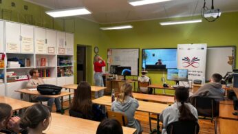 Zdjęcie ukazuje grupę uczniów siedzących w ławkach i patrzących w stronę tablicy multimedialnej, na której wyświetlane jest połączenie wideo. W centralnej części sali dwoje uczniów korzysta z zestawów VR, a nauczycielka w czerwonej bluzce czuwa nad przebiegiem lekcji. Klasa jest jasna, z zielonymi ścianami i nowoczesnym oświetleniem sufitowym. Na regale po lewej stronie widać liczne segregatory i pomoce dydaktyczne.