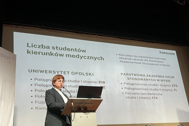 Kobieta w okularach prezentuje dane przy mównicy na tle slajdu zatytułowanego „Liczba studentów kierunków medycznych”. Prezentacja zawiera statystyki dla Uniwersytetu Opolskiego oraz Państwowej Akademii Nauk Stosowanych w Nysie. Sala jest oświetlona światłem scenicznym, a na dole widoczne są fragmenty zielonych foteli.