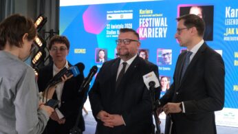 Trzy osoby w formalnych strojach stoją na tle jasnego ekranu z napisem „Festiwal Kariery” i logotypami unijnymi. Mężczyzna w centrum oraz kobieta i mężczyzna po bokach udzielają wywiadu dziennikarce trzymającej mikrofony różnych stacji radiowych i telewizyjnych. W tle widoczne są profesjonalne reflektory estradowe.