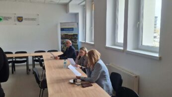 Trzy osoby siedzą przy długim stole w jasnej sali konferencyjnej i analizują dokumenty. Na ścianie widnieją logotypy Wojewódzkiego Urzędu Pracy w Opolu oraz samorządu województwa. W tle widać wysoki stojak reklamowy z informacjami o Funduszach Europejskich.