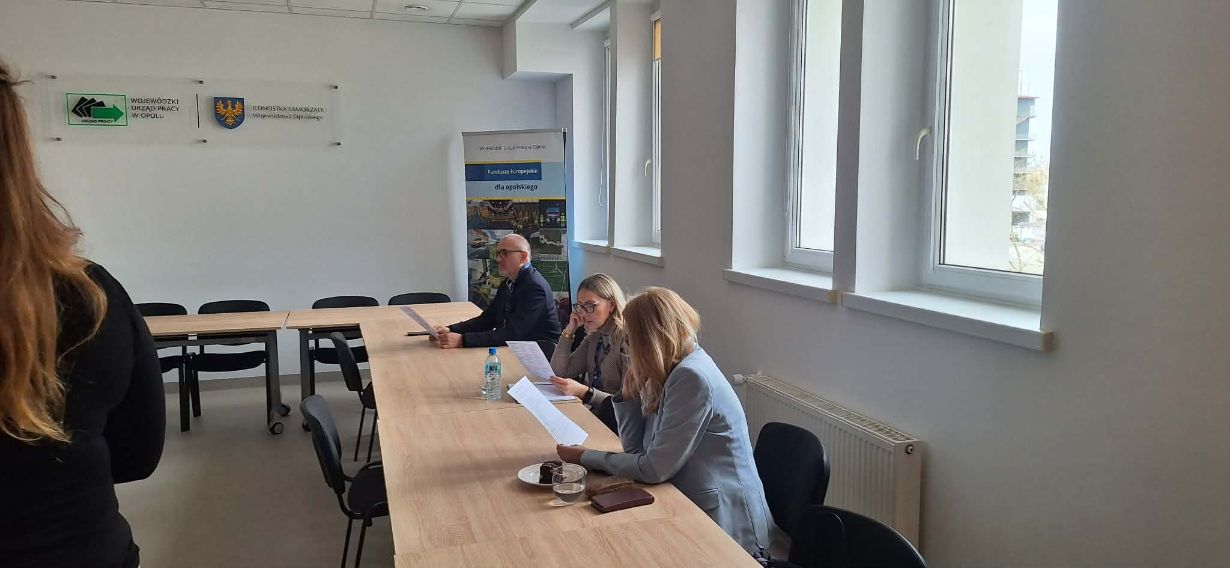 Trzy osoby siedzą przy długim stole w jasnej sali konferencyjnej i analizują dokumenty. Na ścianie widnieją logotypy Wojewódzkiego Urzędu Pracy w Opolu oraz samorządu województwa. W tle widać wysoki stojak reklamowy z informacjami o Funduszach Europejskich.