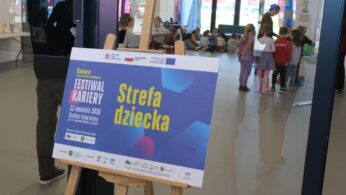 Tablica informacyjna na drewnianej sztaludze z napisem „Festiwal Kariery – Strefa dziecka” oraz datą 22 kwietnia 2026 r. W tle, za szklanymi drzwiami, widać rozmyte sylwetki dzieci bawiących się pod opieką dorosłych w jasnym korytarzu. Na dole plakatu umieszczono liczne logotypy partnerów i organizatorów wydarzenia.