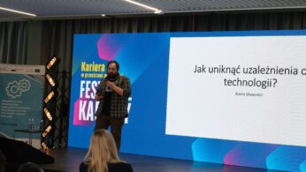 Mężczyzna z brodą w koszuli w kratę stoi na scenie i prowadzi prezentację. Za nim znajduje się wielki ekran z napisem „Jak uniknąć uzależnienia od technologii?” oraz nazwiskiem prelegenta. Z przodu widoczny jest tył głowy jednej z osób siedzących na widowni.