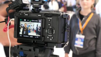Zbliżenie na tył profesjonalnej kamery filmowej zamontowanej na statywie, na której ekranie widać podgląd nagrywanej dziewczyny. W tle, poza ostrością, stoi ta sama dziewczyna z identyfikatorem na szyi, przygotowana do nagrania. Na kamerze widoczne są akcesoria marki Neewer oraz kontrolki statusu nagrywania.