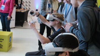 Grupa nastolatków testuje gogle do wirtualnej rzeczywistości (VR) w strefie warsztatowej. Chłopak w czarnej koszulce ma założony zestaw VR i trzyma kontrolery, a jego koledzy obok przyglądają się temu z uśmiechem. W tle widać inne osoby oraz stoiska z logotypami uczelni wyższych.