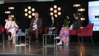 Widok na salę prelekcyjną, gdzie na scenie występuje ekspert prowadzący wykład. Prelegent gestykuluje, omawiając slajdy wyświetlane na dużym ekranie multimedialnym. Słuchacze siedzą w rzędach, a niektórzy z nich robią notatki lub zdjęcia prezentacji.