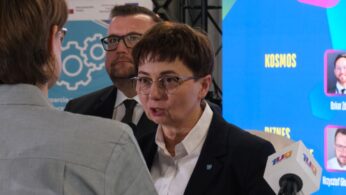 Bliskie ujęcie kobiety w okularach i ciemnej marynarce, która z zaangażowaniem wypowiada się do mikrofonu podczas wywiadu. Za nią stoi mężczyzna w okularach, a w tle widać fragment kolorowej grafiki konferencyjnej. Kobieta ma wpięty w klapę mały herb województwa opolskiego.