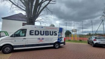 Biały samochód dostawczy z dużym napisem „EDUBUS” i hasłem „Nauka przez całe życie” zaparkowany na brukowanym placu przed szkołą. Pojazd ozdobiony jest kolorową grafiką przedstawiającą robota oraz logotypami projektowymi na tle pochmurnego nieba i dużego drzewa bez liści. W tle widać boisko do koszykówki ogrodzone wysoką siatką oraz zaparkowane obok auta osobowe.