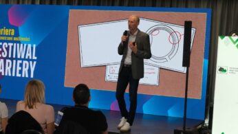 Ujęcie panelu dyskusyjnego, w którym bierze udział czworo ekspertów siedzących w wygodnych fotelach na scenie. Przed nimi ustawione są stoliki z wodą, a każdy z uczestników trzyma mikrofon. Nad panelistami znajduje się ekran z tematem debaty i nazwiskami prelegentów.