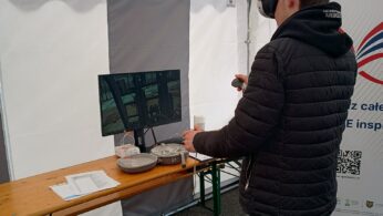 Osoba ubrana w czarną kurtkę, dżinsy i zestaw VR używa ręcznych kontrolerów w białym namiocie. Stoi przy drewnianym stole z monitorem, sprzętem VR i dokumentami. Pionowy znak z kolorową grafiką i tekstem jest częściowo widoczny obok nich.