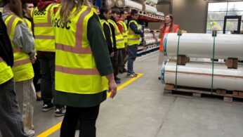 Grupa osób w żółtych kamizelkach ochronnych stoi w magazynie i słucha osoby w pomarańczowej kamizelce. Rolki materiału są ułożone na regałach i paletach. Magazyn ma wysokie sufity, świetliki i oświetlenie przemysłowe.