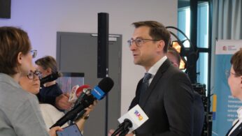 Profil mężczyzny w okularach i ciemnym garniturze, który mówi do grupy dziennikarzy trzymających mikrofony i dyktafony. Akcja dzieje się w nowoczesnym wnętrzu z widocznym w tle sprzętem oświetleniowym. Mężczyzna ma pogodny wyraz twarzy i gestykuluje podczas wypowiedzi.