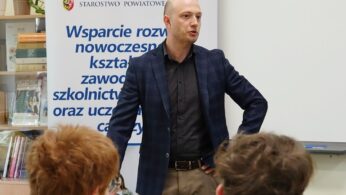 Zbliżenie na prelegenta w granatowej marynarce w kratę i brązowych spodniach, który przemawia, opierając ręce na biodrach. Stoi obok baneru reklamowego z logo 