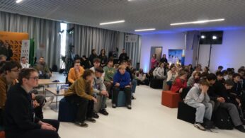Szerokie ujęcie sali konferencyjnej wypełnionej młodzieżą siedzącą na kolorowych pufach. Uczestnicy z uwagą patrzą w stronę niewidocznej na zdjęciu sceny, a niektórzy mają na szyjach identyfikatory na smyczach. W tle widać nowoczesne oświetlenie sufitowe oraz profesjonalne statywy z kamerami i lampami.