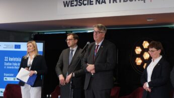 1 (6).jpg: Zbliżenie na dłonie osób siedzących przy stoliku, które wypełniają dokumenty lub ankiety podczas wydarzenia. Na stole leżą ulotki promocyjne, długopisy oraz identyfikatory na smyczach. W tle widać rozmyte sylwetki innych uczestników festiwalu.