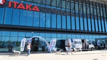 Kadr z wydarzenia przedstawiający osoby gromadzące się wokół stoisk informacyjnych w jasnym, nowoczesnym obiekcie sportowym. Na pierwszym planie widać fragmenty ścianek wystawienniczych oraz uczestników prowadzących rozmowy. Przez duże przeszklenia w tle przebija widok na trybuny stadionu i otwartą przestrzeń boiska.
