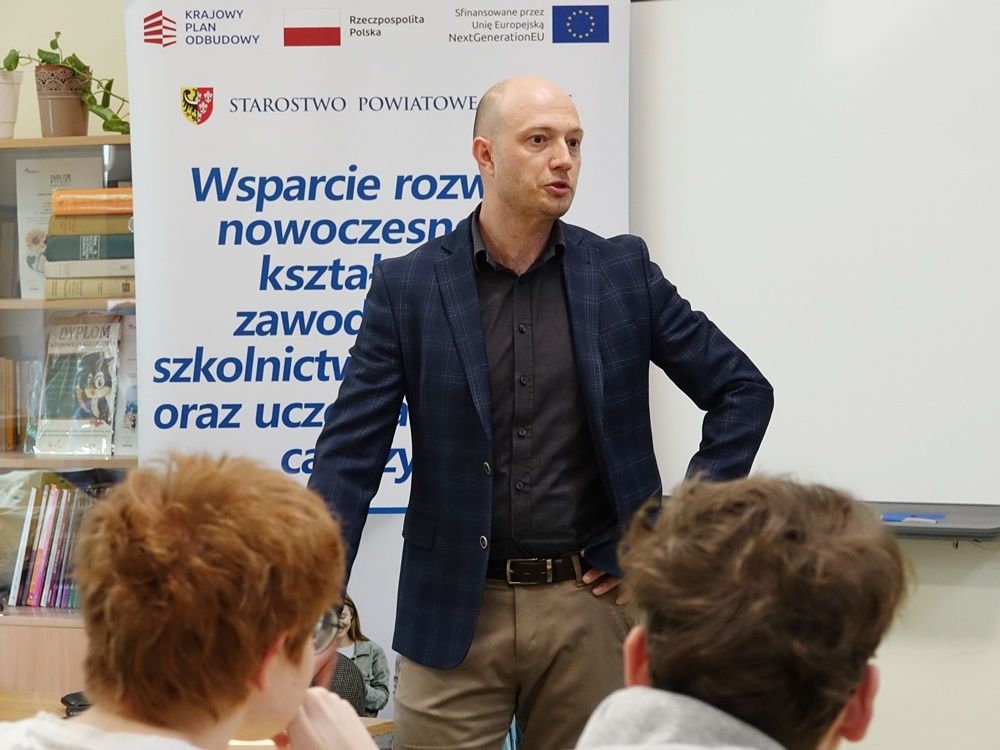 Zbliżenie na prelegenta w granatowej marynarce w kratę i brązowych spodniach, który przemawia, opierając ręce na biodrach. Stoi obok baneru reklamowego z logo 