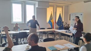 Grupa osób uczestniczy w warsztatach przy stołach ustawionych w podkowę, a jedna z kobiet stoi i rozdaje materiały. W tle znajdują się flagi Polski, Unii Europejskiej oraz województwa opolskiego. Na biurkach leżą niebieskie notesy i identyfikatory na smyczach.