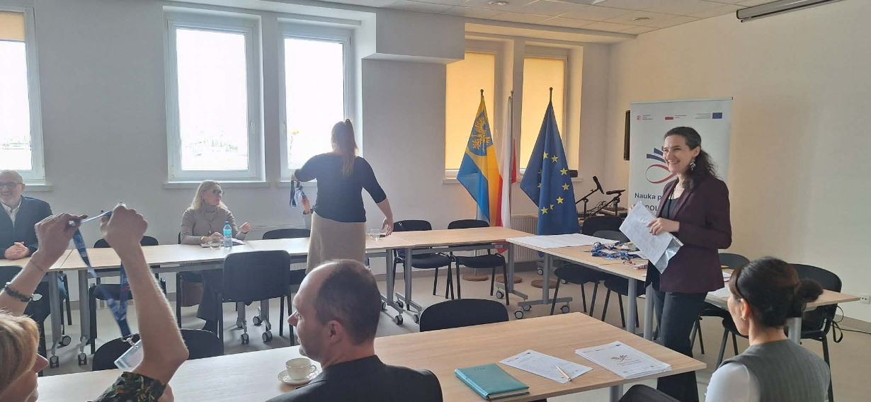 Grupa osób uczestniczy w warsztatach przy stołach ustawionych w podkowę, a jedna z kobiet stoi i rozdaje materiały. W tle znajdują się flagi Polski, Unii Europejskiej oraz województwa opolskiego. Na biurkach leżą niebieskie notesy i identyfikatory na smyczach.