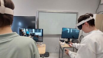 Zbliżenie na dwóch uczniów zanurzonych w rzeczywistości wirtualnej przy użyciu gogli VR i kontrolerów ręcznych. Na stojących przed nimi monitorach wyświetlany jest podgląd obrazu z gogli, przedstawiający wnętrze budynku oraz techniczne pomieszczenie. Stanowiska są wyposażone w laptopy i gogle połączone przewodami, a w tle widać czystą, białą tablicę szkolną.