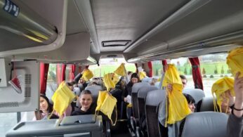 Wnętrze autokaru wypełnione uśmiechniętą młodzieżą, która pozuje do zdjęcia. Pasażerowie trzymają w górze żółte worki turystyczne, tworząc radosny i zintegrowany widok. Korytarz pojazdu jest czysty, a za oknami widać zielony krajobraz.