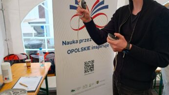 Mężczyzna w czarnej kurtce korzysta z białych słuchawek wirtualnej rzeczywistości i kontrolerów pod namiotem. Za nim znajduje się baner z polskim tekstem i logo, nawiązujący do nauki i Opola. W pobliżu na drewnianych stołach leżą papiery, butelki i szary futerał na sprzęt VR.