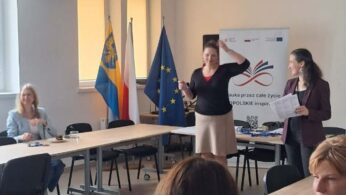 Uczestnicy spotkania siedzą przy stole, podczas gdy dwie prowadzące stoją przy banerze z napisem „Nauka przez całe życie – OPOLSKIE inspiruje”. Na stole widać filiżanki z kawą, wodę oraz rozłożone materiały szkoleniowe. Sala jest dobrze oświetlona światłem dziennym wpadającym przez duże okna.