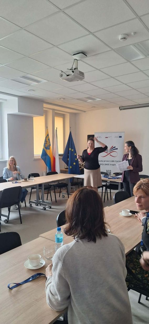 Uczestnicy spotkania siedzą przy stole, podczas gdy dwie prowadzące stoją przy banerze z napisem „Nauka przez całe życie – OPOLSKIE inspiruje”. Na stole widać filiżanki z kawą, wodę oraz rozłożone materiały szkoleniowe. Sala jest dobrze oświetlona światłem dziennym wpadającym przez duże okna.