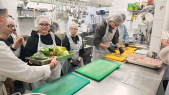 Grupa uczniów w fartuchach ochronnych i czepkach uczestniczy w zajęciach praktycznych w profesjonalnej kuchni. Jeden z uczniów kroi mięso na żółtej desce, podczas gdy inni obserwują proces lub trzymają tacę z warzywami. W tle widoczne jest lśniące wyposażenie gastronomiczne ze stali nierdzewnej.