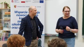 Zdjęcie przedstawia dwoje prelegentów – mężczyznę w marynarce i kobietę w ciemnej bluzce – przemawiających do grupy uczniów w klasie. Prelegenci stoją obok baneru promocyjnego 